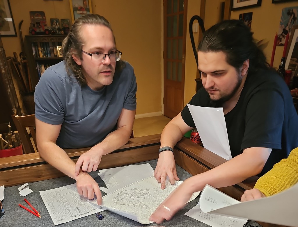 Two men are gathered around a table covered in maps and papers, discussing strategy or planning. One man, wearing glasses and a blue shirt, is pointing at a map while the other man, in a black shirt, is looking intently at the documents. There are also drinks and stationery visible on the table.