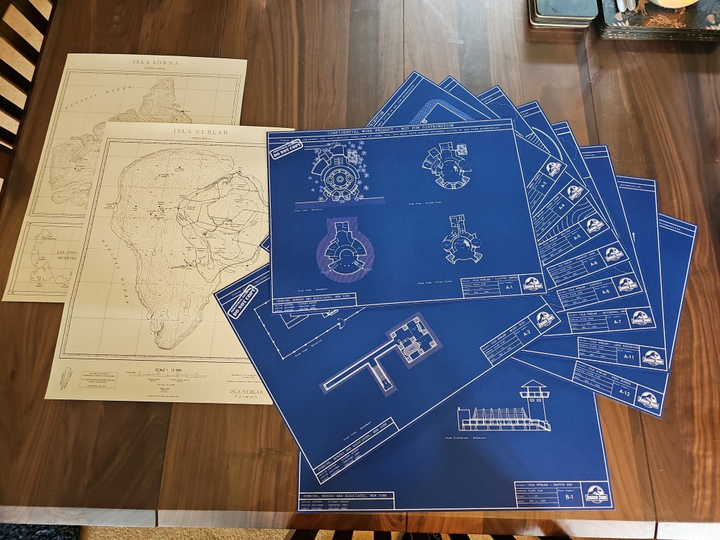 A collection of architectural blueprints and maps laid out on a wooden table, including detailed plans for Isla Nublar and Isla Sorna.
